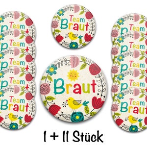 Könnte beinhalten: Zwölf runde, floral gemusterte Knöpfe mit dem Text "Team Braut" und einem gelben Vogel. Ein größerer Knopf mit einer Sonne, Blumen und dem Text "Braut" mit einem gelben Vogel. Der Text "1 + 11 Stück" befindet sich am unteren Rand des Bildes.