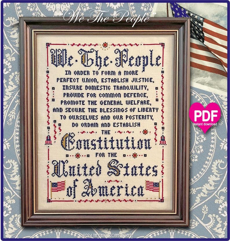 May include: A framed cross-stitch sampler on light beige fabric featuring the Preamble to the United States Constitution in dark blue and red lettering. The text is surrounded by a decorative red border and two small American flag illustrations.