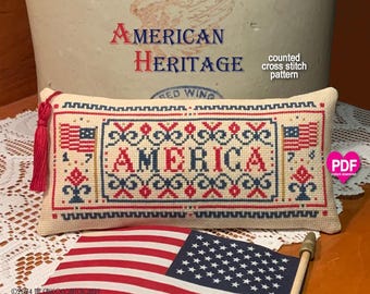 May include: A rectangular cross stitch sampler on cream fabric featuring the word AMERICA in red and blue, flanked by two small American flags. The design includes decorative borders, red and blue floral motifs, and a red tassel on the left side.