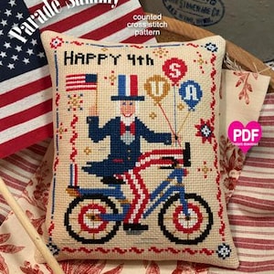Könnte beinhalten: Ein Zählmuster für Kreuzstich für ein Kissen mit einem Cartoon-Onkel Sam, der ein Fahrrad fährt, mit den Worten "HAPPY 4th" darüber. Das Fahrrad hat rot-weiß-blaue Reifen und die Onkel-Sam-Figur trägt eine blaue Jacke, ein weißes Hemd und rot-weiß-blaue gestreifte Hosen. Das Muster heißt "Parade Sammy".
