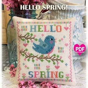 Puede incluir: Patrón de punto de cruz con un pájaro azul en una rama con flores rosas y el texto "HELLO SPRING!" en letras de colores. El patrón está sobre un fondo blanco y está enmarcado por una almohada blanca.