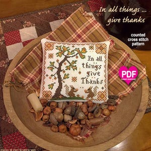 以下が含まれることがあります： 秋の葉がついた木、2匹のリス、そして「In all things give thanks」という文字が刺繍された正方形のカウントクロスステッチパターン。オレンジ、茶色、緑の色合いで構成されており、PDFダウンロードファイルとして提供されています。