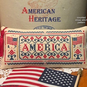 Op de afbeelding: Geteld kruissteekpatroon voor een kussen met het woord "AMERICA" in rood, wit en blauw. Het patroon bevat een kleine Amerikaanse vlag aan elke kant van het woord. Het patroon heet "American Heritage".