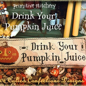 May include: A cross-stitch sampler with a pumpkin and the text "Drink Your Pumpkin Juice". The sampler is framed and surrounded by fall-themed decorations, including candles, a small doll, and dried leaves.