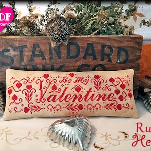 May include: A rectangular, beige cross-stitch pillow with the words "Be My Valentine" in red, surrounded by hearts and floral designs. The image includes a wooden box, metal heart-shaped molds, and a lit red candle.