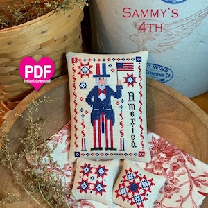 Könnte beinhalten: Ein rechteckiger Kreuzstich-Sampler mit Uncle Sam, der eine amerikanische Flagge hält, mit dem Text America, begleitet von zwei kleineren quadratischen Kreuzstichkissen mit rot-weiß-blauen geometrischen Sternmustern auf cremefarbenem Stoff.
