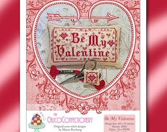 May include: A cross stitch pattern for a pillow with the text "Be My Valentine" in red lettering on a white background. The pattern is surrounded by a red heart-shaped frame with floral details. The pattern is 300 x 55 stitches and uses DMC threads.