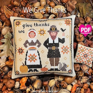 May include: A square cross-stitch cushion featuring a person in a bonnet and a person in a hat with the text "give thanks we". A smaller matching square cushion with a geometric star pattern sits to the right. Both items are beige with black, orange, and brown thread details.