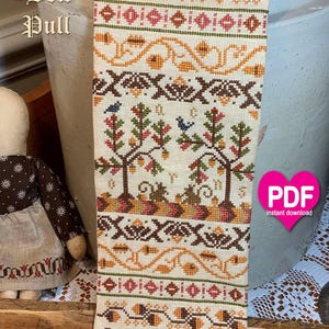 May include: A vertical cross-stitch bell pull on cream fabric featuring repeating patterns of brown acorns, orange leaves, and small blue birds in trees. The text Acorn Bell Pull is printed on the left. The piece is finished with small metal beads.