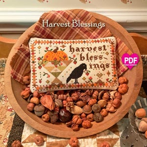 May include: A rectangular cross-stitch sampler featuring the text Harvest Blessings, a large orange pumpkin, and a black crow. The design is bordered by small orange and brown geometric patterns on a cream fabric, displayed in a wooden bowl.
