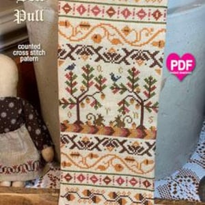 May include: A counted cross stitch pattern for a needlepoint project titled "Acorn Bell Pull". The pattern features a cream-colored fabric with brown, orange, and green embroidery depicting acorns, leaves, and birds.