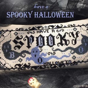 May include: A rectangular counted cross stitch pattern on cream fabric featuring the text "HAVE A SPOOKY HALLOWEEN" in black and blue gothic lettering, surrounded by black and blue ghost illustrations and a decorative border, with a pink PDF icon.