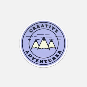 May include: A purple circle sticker with a white outline. The sticker has the text "CREATIVE ADVENTURER" in black text around the edge. Inside the circle is a graphic of three mountains that look like pencils. The mountains are yellow with black tips and are set against a light blue background.