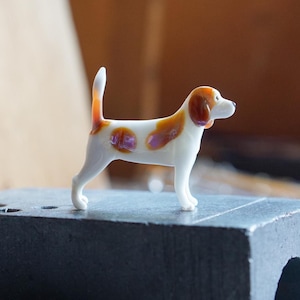 Könnte beinhalten: Eine kleine Glasfigur eines Hundes im Profil. Der Hund ist überwiegend weiß mit braunen Flecken auf Rücken, Ohren und Schwanz. Er hat eine glatte, glänzende Oberfläche und ist in stehender Position mit aufgerichtetem Schwanz dargestellt.