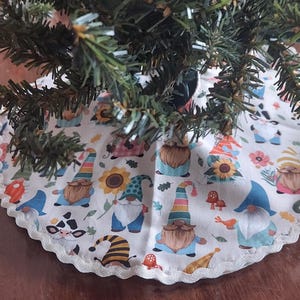 May include: A white Christmas tree skirt with a scalloped edge, decorated with colorful gnomes, sunflowers, and mushrooms. The skirt is placed around the base of a green Christmas tree.