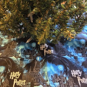 May include: A Christmas tree skirt with a black and blue design featuring the text "Harry Potter" and illustrations of a castle. The skirt has a silver lace trim and is placed under a green Christmas tree with white lights.