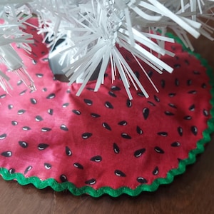 May include: A red Christmas tree skirt with a watermelon design, featuring black seeds and a green scalloped edge. The skirt is under a white Christmas tree, creating a festive summer holiday theme.