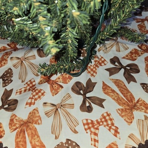 May include: A circular fabric tree skirt featuring a repeating pattern of various bows in shades of orange, brown, and cream. The bows include striped, checkered, floral, and polka dot designs. The edge is finished with a dark brown rickrack trim.