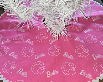 May include: A circular pink tree skirt featuring a repeating white line-art pattern of the Barbie silhouette logo and the Barbie script text. The edge of the fabric is finished with a white rickrack trim, and a white artificial tree is centered.