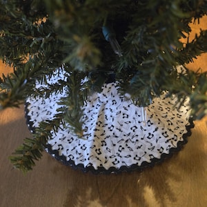 May include: A white Christmas tree skirt with a black musical note pattern and a scalloped black trim. The skirt is placed around the base of a green artificial Christmas tree.