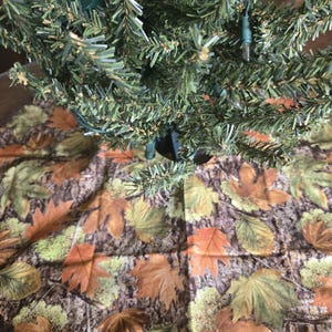 May include: A Christmas tree skirt with a camouflage pattern of brown, green, and orange leaves. The skirt has a brown scalloped edge and is placed under a green artificial Christmas tree with lights.