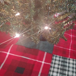 May include: A red and white plaid Christmas tree skirt with a white scalloped edge. The skirt is made of fabric and has a patchwork design. The skirt is decorated with a green and white plaid pattern.