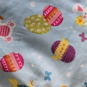 May include: Light blue fabric with a repeating Easter-themed pattern. The design includes white bunnies, colorful Easter eggs, and small butterflies. The eggs are decorated with stripes and polka dots.