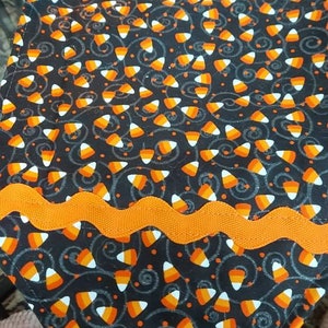 May include: Black table runner with an orange and white candy corn pattern. The runner has an orange scalloped trim.