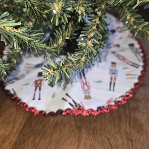 May include: A white Christmas tree skirt with a red scalloped edge. The skirt features a repeating pattern of toy soldiers in various poses. The tree skirt is under a green Christmas tree.