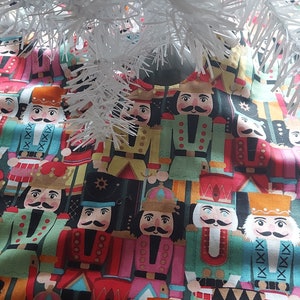 May include: A red and white Christmas tree skirt with a nutcracker pattern. The skirt has a scalloped edge and is made of fabric.