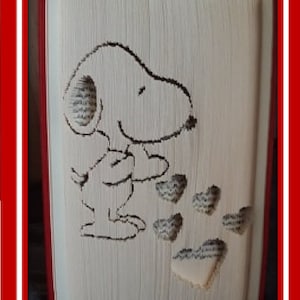 May include: A folded book art piece featuring Snoopy holding hearts. The artwork is created from the pages of a book, with the character and hearts outlined in black. The book's edges are red, and the text "Livres pliés de Kiki / Book folds by Kiki" is at the top.