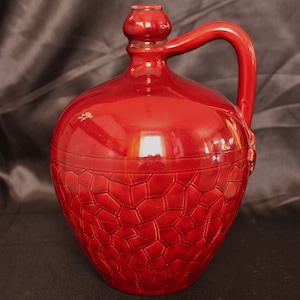May include: A red ceramic jug with a textured surface and a handle. The jug has a rounded body and a small spout.