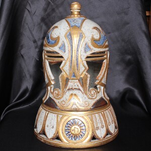 May include: A white ceramic helmet with gold and blue accents. The helmet has a decorative design and a round base.