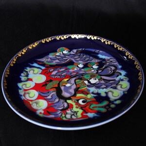 Peut inclure: Une assiette en céramique bleue avec un bord doré et un motif coloré d'un dragon avec des oiseaux et des nuages. Le dragon est rouge, violet et vert, et les oiseaux sont jaunes et bleus.