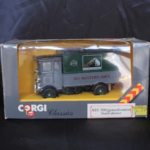 May include: A gray Corgi Classics diecast model of an AEC 508 forward control 5-ton cabover truck. The truck has a green box body with the words "His Master's Voice" and a logo of a dog listening to a gramophone. The box is labeled "Playworld" and has a price of $10.99.