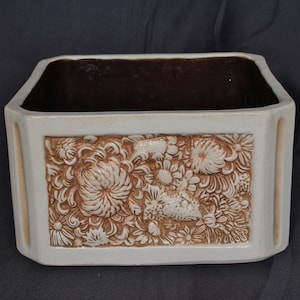 Può includere: Un vaso di ceramica bianca con interno smaltato marrone. Il vaso ha un design floreale in rilievo con fiori e foglie marroni.