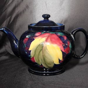Peut inclure: Une théière bleue avec un motif floral composé de feuilles rouges, jaunes et vertes et de baies violettes. La théière a un corps rond et un couvercle.