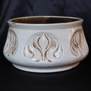 May include: White ceramic planter with a brown glaze on the inside rim. The planter has a rounded shape and features a repeating pattern of stylized floral designs.