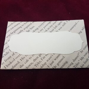 May include: A white envelope with a blank, white, rounded rectangular label. The envelope is made of paper and has a textured surface. The label is adhered to the envelope and has a slightly raised edge.