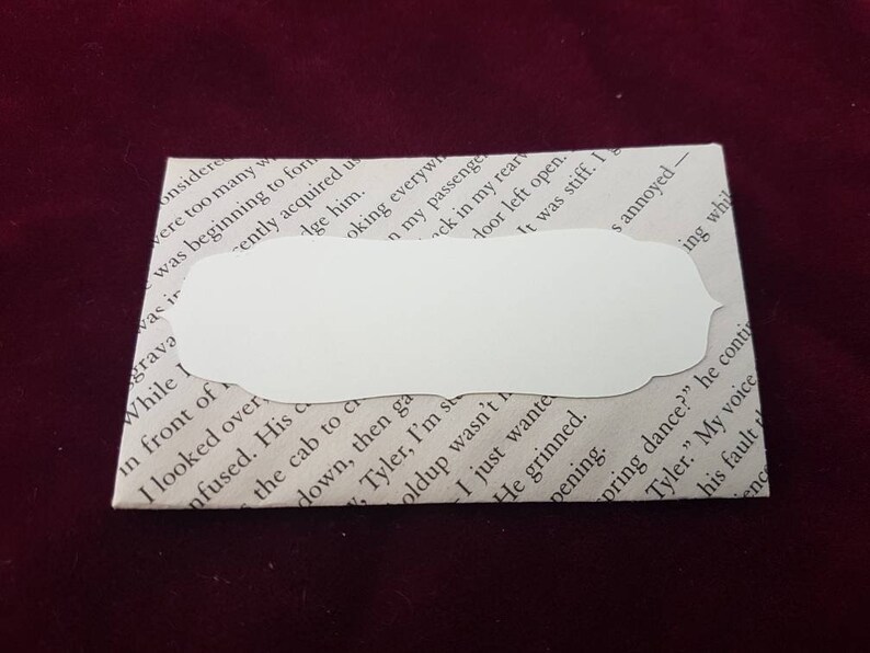 May include: A white envelope with a blank, white, rounded rectangular label. The envelope is made of paper and has a textured surface. The label is adhered to the envelope and has a slightly raised edge.
