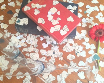 May include: Two clear glass champagne flutes, a red hardcover book, and a dark blue hardcover dictionary are surrounded by numerous small heart-shaped cutouts made from printed book pages. A single bright red gerbera daisy lies nearby.