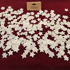 May include: A large quantity of white paper stars scattered on a red surface. The stars are made of paper and have a printed design. The text "[5]0[0]" is visible on a small brown tag.