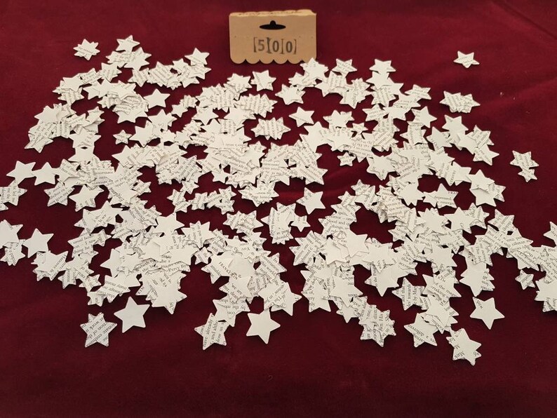 May include: A large quantity of white paper stars scattered on a red surface. The stars are made of paper and have a printed design. The text "[5]0[0]" is visible on a small brown tag.