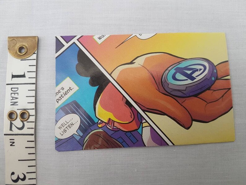 May include: A comic book panel featuring a hand holding a blue and silver disc with the letter "A" in the center. The text "Well... listen..." is in a speech bubble.