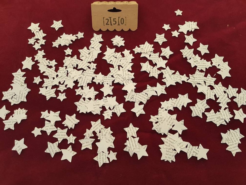 May include: A scattering of white paper stars on a red background. The stars are made of paper with printed text. The text on the stars is not legible. There is a small brown tag with the text "[2]5[0]" in the upper center of the image.