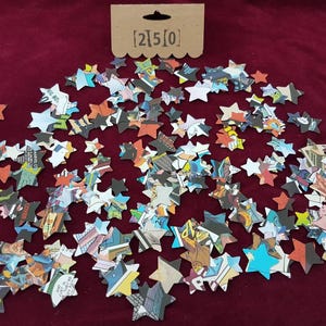 May include: A scattering of colorful star-shaped confetti made from recycled comic book pages. The confetti is on a red background. The text [2]5[0] is printed on a brown tag.