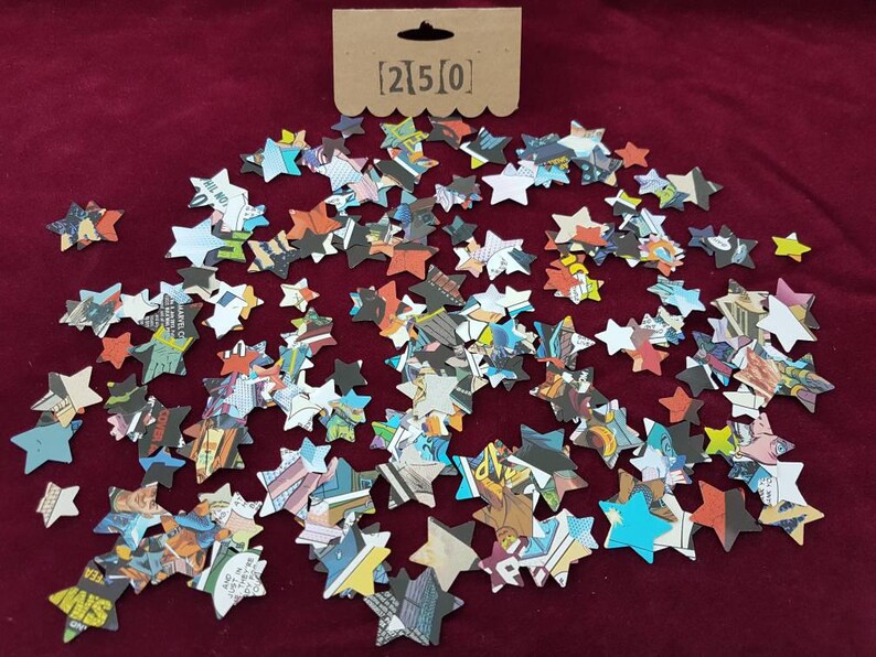 May include: A scattering of colorful star-shaped confetti made from recycled comic book pages. The confetti is on a red background. The text [2]5[0] is printed on a brown tag.