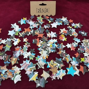 May include: A scattering of colourful star-shaped confetti made from recycled comic book pages. The confetti is on a red background. The text [2]5[0] is printed on a brown tag.