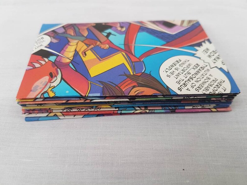 May include: A stack of colorful comic book pages featuring a superhero character with a red, yellow, and blue costume. The text on the pages reads "IT THEROP PROBABLY A BONKERS MUTATION OF A TYRANNOSAURUS REX...BUT THE IMPORTANT THING IS HE'S FRIENDLY. WHY YOU BOY AWAY FROM ME".