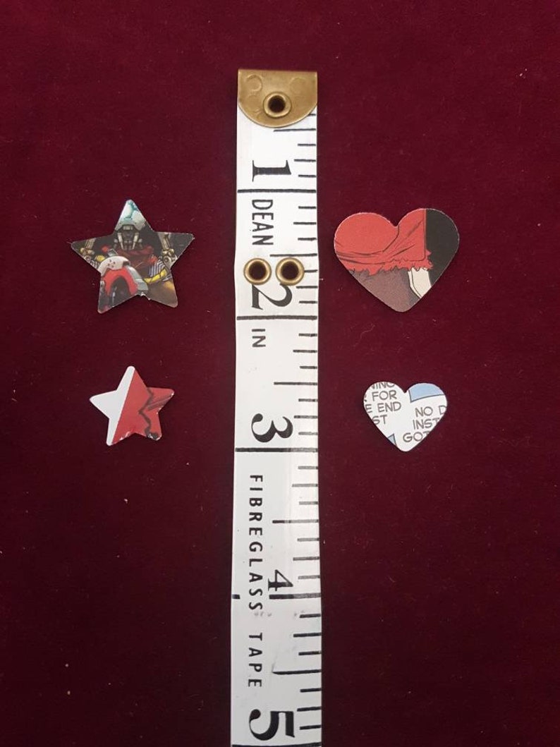 May include: Three paper cutouts of a star, a heart, and a star with a red, white and blue design. The cutouts are placed on a red surface next to a white measuring tape with black markings. The tape measures in inches and is labelled "Dean" and "Fibreglass Tape".